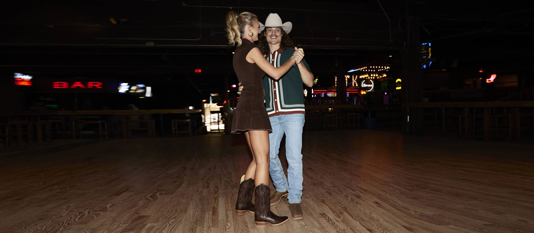 A blonde woman with her hair in a ponytail looking away from the camera and dancing with a man with long brown curly hair.